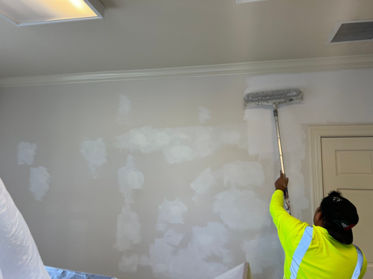 Painter rolling an office wall while furniture stays protected under plastic during an occupied commercial repaint